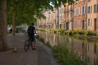 Marseille canal path with a person pausing on a bike, terracotta buildings, wild grasses, and gentle water reflections in soft afternoon light.