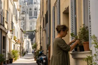 Marseille resident watering plants by a window on a quiet street with Cathédrale de la Major's facade softly blurred in the background, under gentle Mediterranean light.