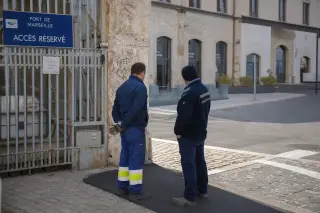 Two Marseille dock workers talking near old port warehouses with visible layers of urban renewal, under soft Mediterranean daylight.