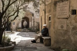 Elderly local resting on a bench in the sunlight-dappled courtyard of a historic Marseille convent with textured stone walls and discreet architectural details.