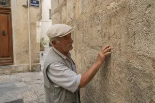 Elderly man quietly touching a weathered medieval wall in a narrow Marseille alley, with subtle daylight and an authentic, lived-in atmosphere.