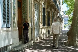 Two local residents studying architectural details while strolling along a shaded Marseille street lined with ornate XIXth-century facades and sun-dappled walls.