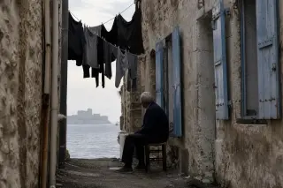 Seaside Marseille alley with weathered walls, lone local seated under laundry lines, distant Château d’If softened by morning mist.