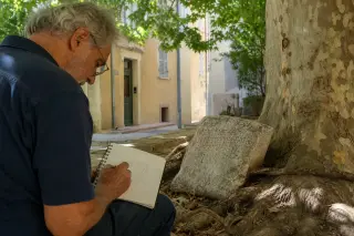 Elderly local sketches ancient Greek-inscribed stone fragment under a leafy tree in a quiet Marseille courtyard, soft natural Mediterranean light, intimate and authentic urban atmosphere.