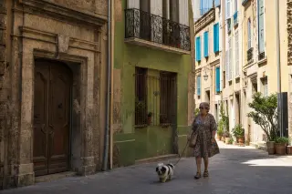 Elderly woman with dog strolling along a narrow, shaded Marseille street, aged colorful buildings with historical details and soft morning light.