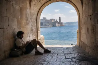 A local quietly sketching in a shaded stone archway overlooking the harbor, with Fort Saint-Nicolas faintly visible across the water under soft daylight.