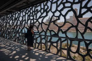 Under the MuCEM's intricate facade, a woman quietly observes Marseille, cast in soft Mediterranean light with old fort stones and modern architecture blending in the background.