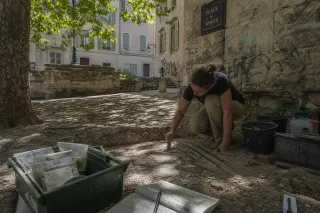 Archaeologist studying a mosaic fragment in a sun-dappled Marseille square, surrounded by ancient stone walls and excavation tools, in a calm and authentic atmosphere.
