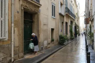 Elderly resident adjusting bag near a weathered 19th-century Marseille doorway on a damp, pastel-colored street